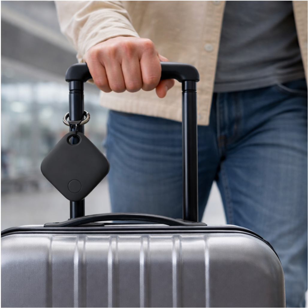 Lifestyle Image with a man holding a suitcase handle with the air tag GPS tracker product (colour back) attached to the suitcase handle, focus on the air tag tracker product in the image, front angle.