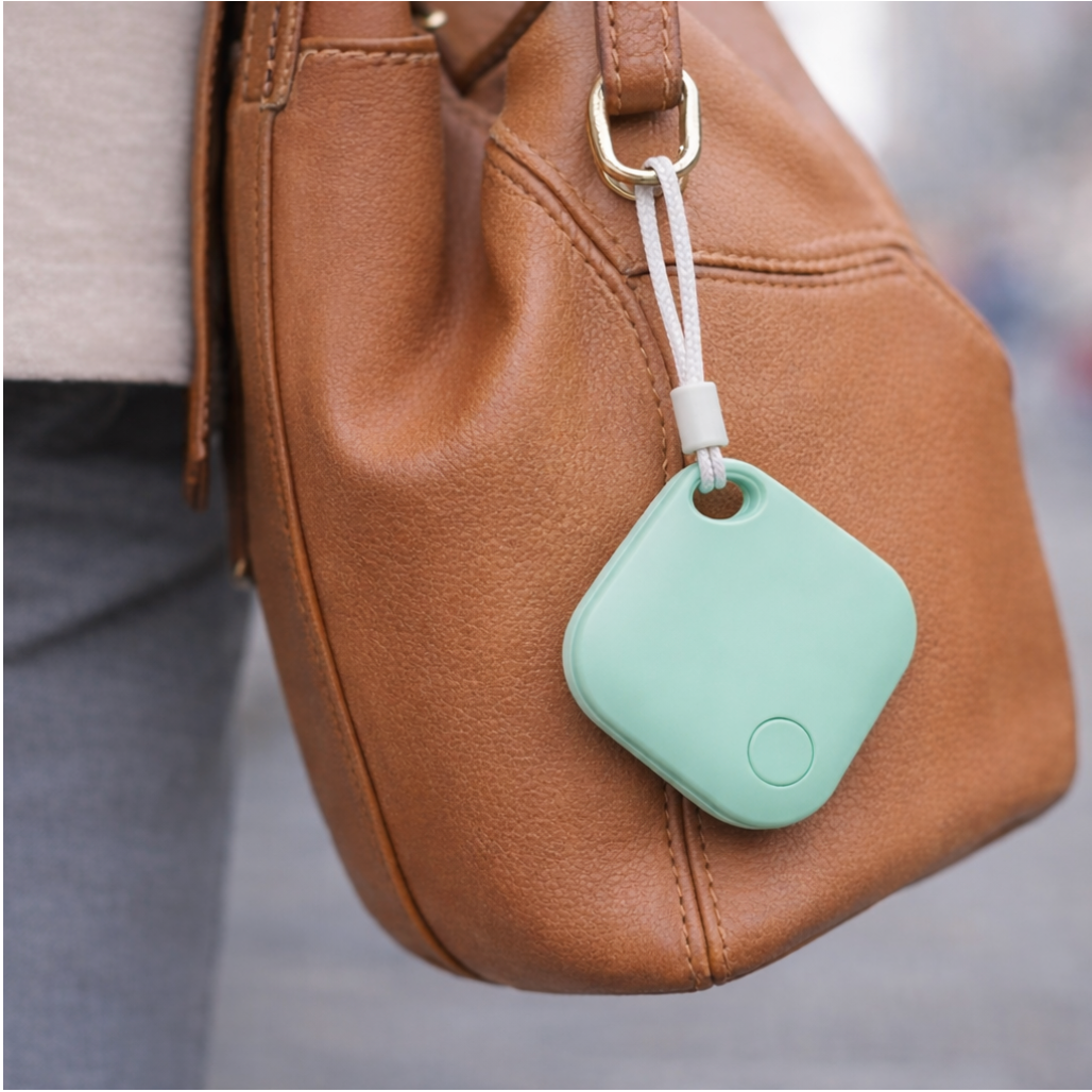 Lifestyle image of a woman's handbag close up with the air tag GPS tracker product (colour white) attached to a gold ring on the bag, focus on the air tag GPS tracker product.